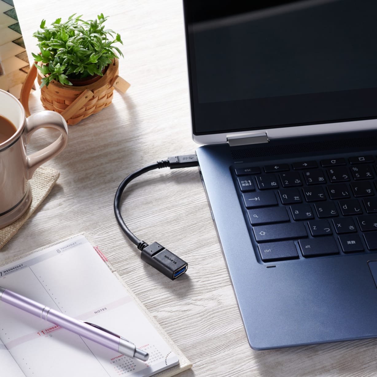 An overhead view of a Chromebook with a USB cable inserted into an open port. A writing pen and notebook lay close by.